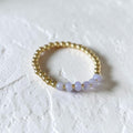 A delicate gold beaded bracelet featuring five small, faceted light purple beads, displayed on a textured white surface.