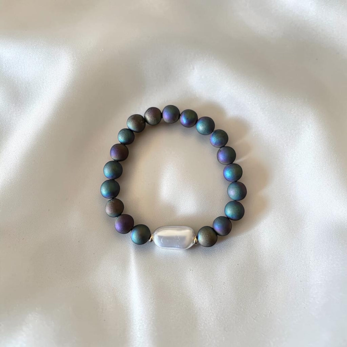 A beaded stretch bracelet made of iridescent dark gray beads with a pearlescent white rectangular accent bead, displayed on a soft white fabric background.