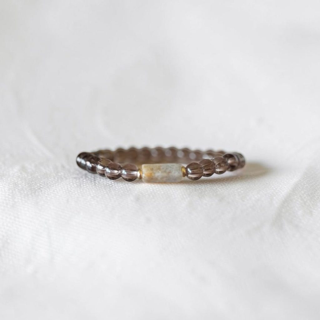 A beaded bracelet with dark brown textured beads and a central rectangular cream-colored stone, resting on a soft white fabric background.
