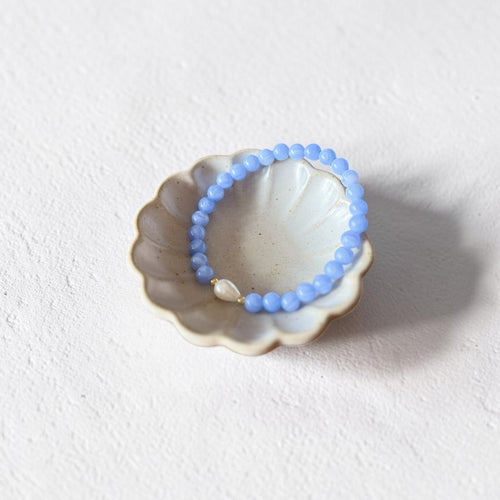 A light blue beaded bracelet with a small white pearl accent, displayed inside a small ceramic shell dish on a textured off-white background.