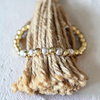 A beaded stretch bracelet with gold, silver, and white beads, wrapped around a natural jute rope tassel display on a light textured background.