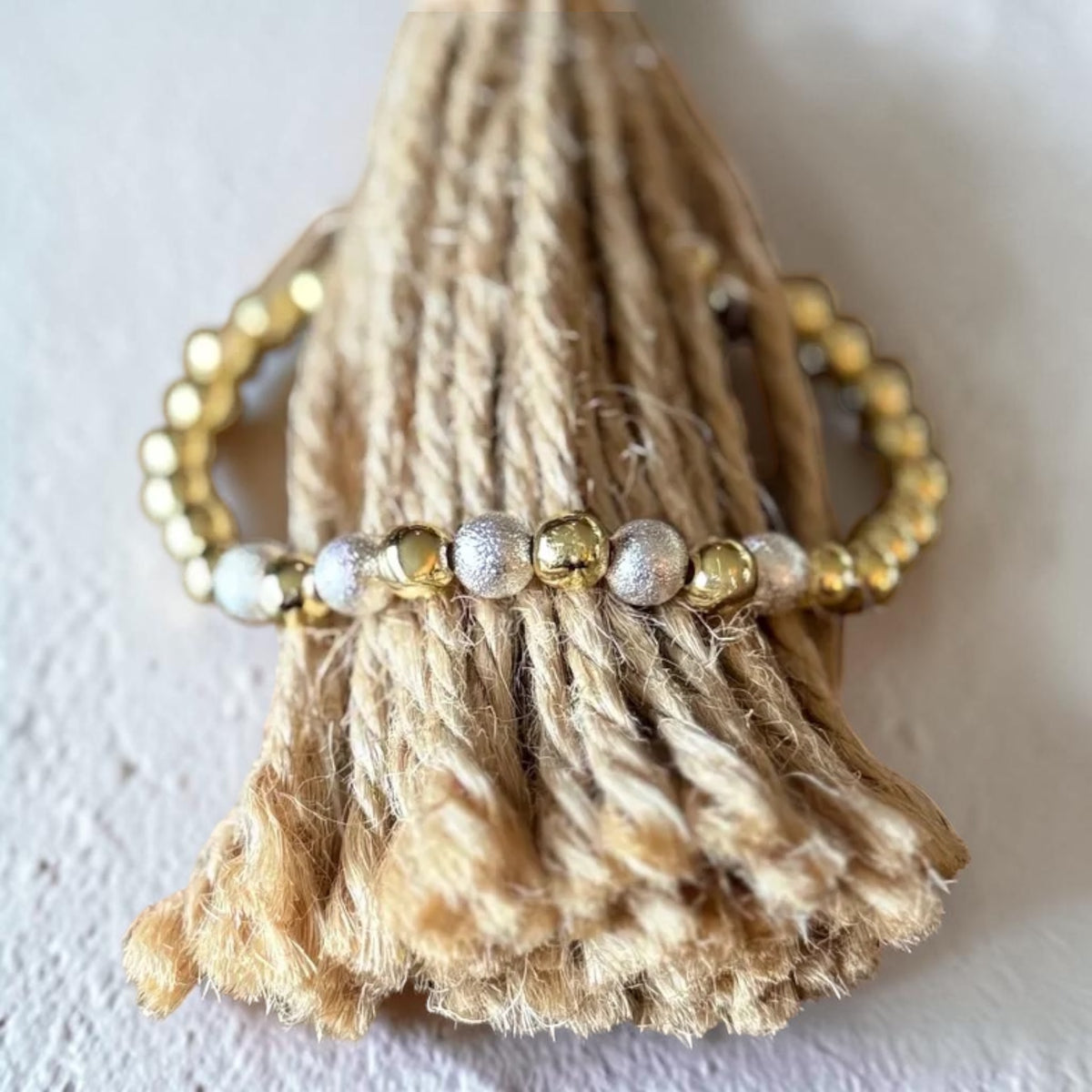 A beaded stretch bracelet with gold, silver, and white beads, wrapped around a natural jute rope tassel display on a light textured background.