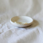 A delicate pearl beaded bracelet displayed inside a small, round, matte off-white ceramic dish on a soft, textured white fabric background.