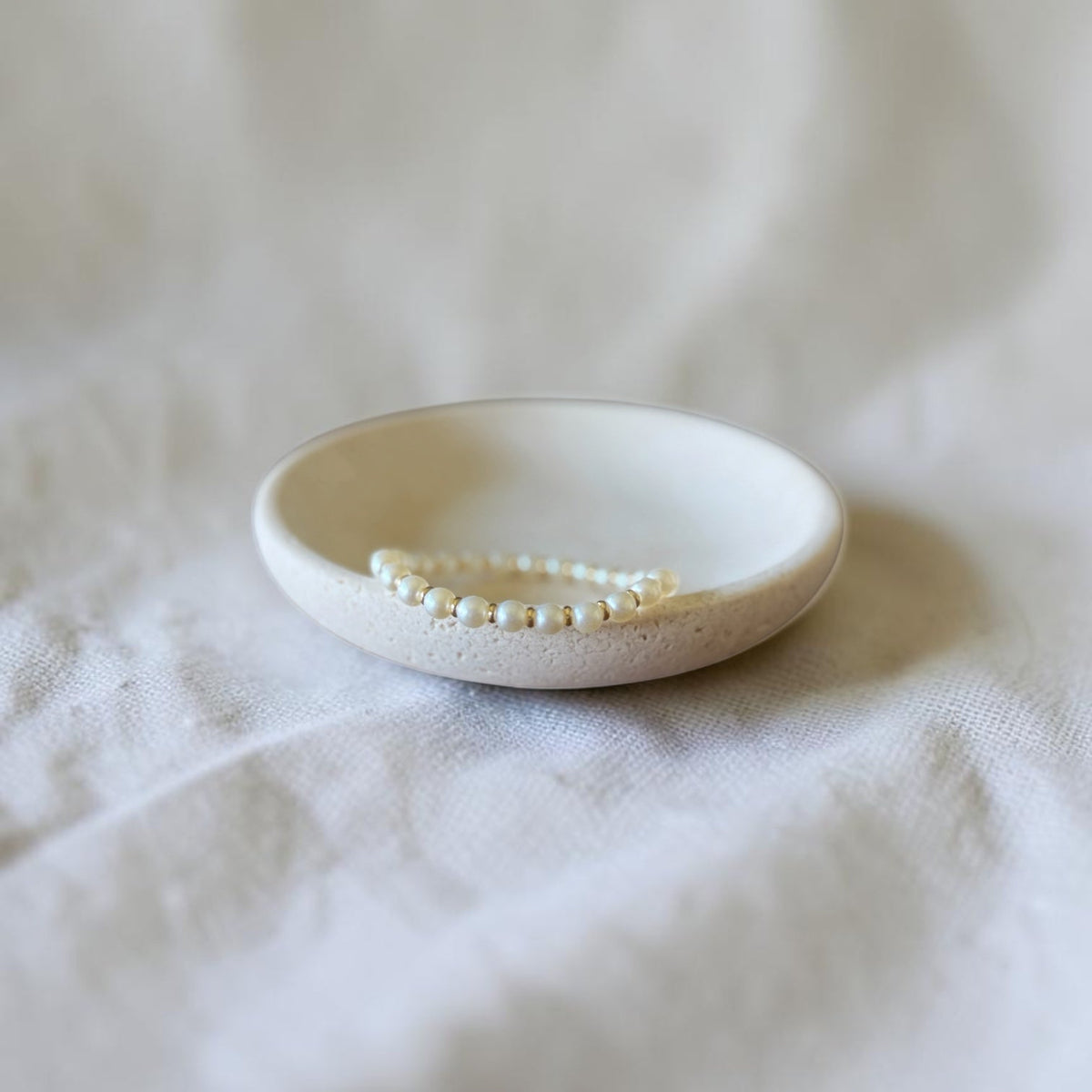 A delicate pearl beaded bracelet displayed inside a small, round, matte off-white ceramic dish on a soft, textured white fabric background.