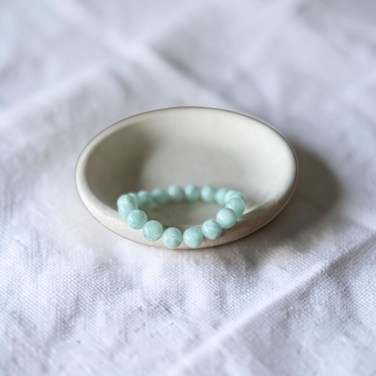 A beaded stretch bracelet made of light mint green beads, displayed inside a small, round, off-white ceramic dish on a textured white fabric background.