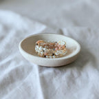 A close-up of a small ceramic dish holding a stack of beaded bracelets with white pearls, peach beads, and small dark accents, set against a soft white fabric background.