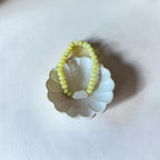 A pale yellow beaded ring with a small white oval stone, displayed on a white scalloped dish against a white background.