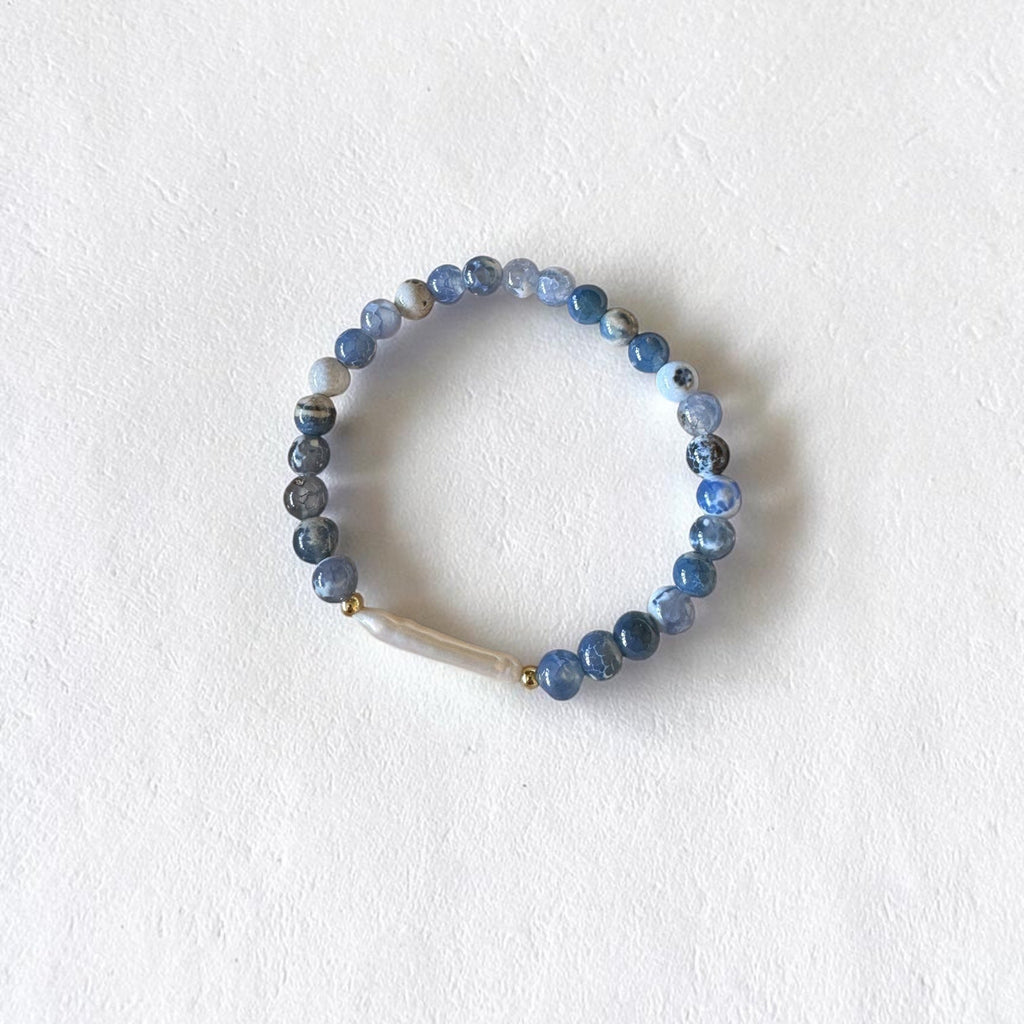 A beaded stretch bracelet featuring round blue and gray marbled beads, a small gold spacer, and a white shell accent, displayed on a textured white background.
