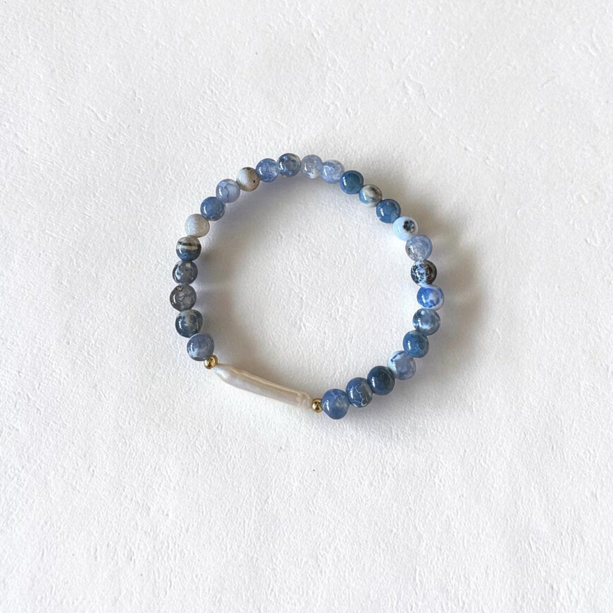 A beaded stretch bracelet featuring round blue and gray marbled beads, a small gold spacer, and a white shell accent, displayed on a textured white background.