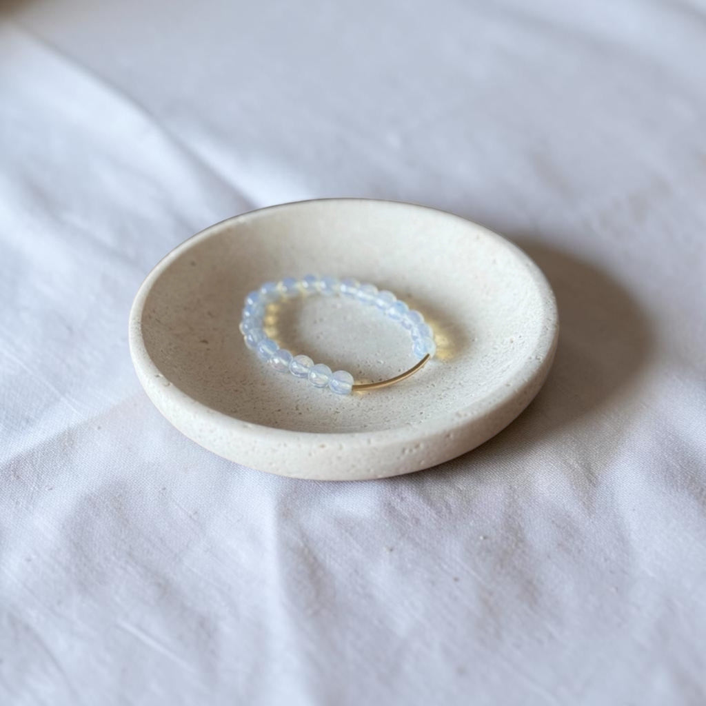A beaded stretch bracelet with pale blue translucent beads and a small gold clasp, displayed inside a round speckled ceramic dish on a white fabric surface.