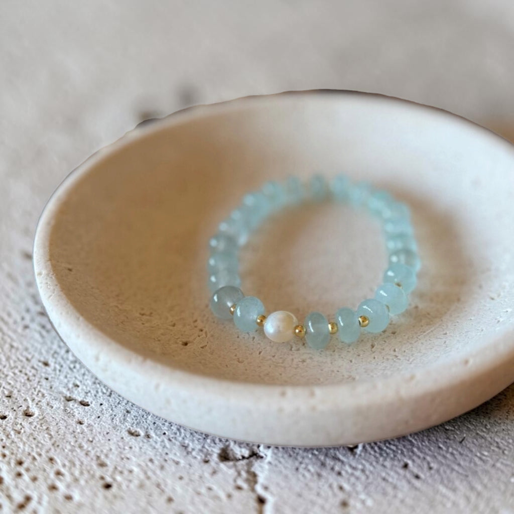 A beaded stretch bracelet with light blue translucent beads, a white pearl-like bead, and small gold spacer beads, displayed inside a shallow beige ceramic dish.