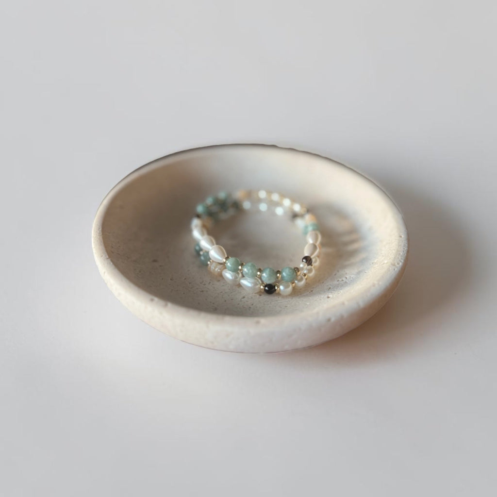 A delicate beaded bracelet with small light green, white, and silver beads resting inside a small, shallow, speckled off-white ceramic dish on a neutral light gray background.
