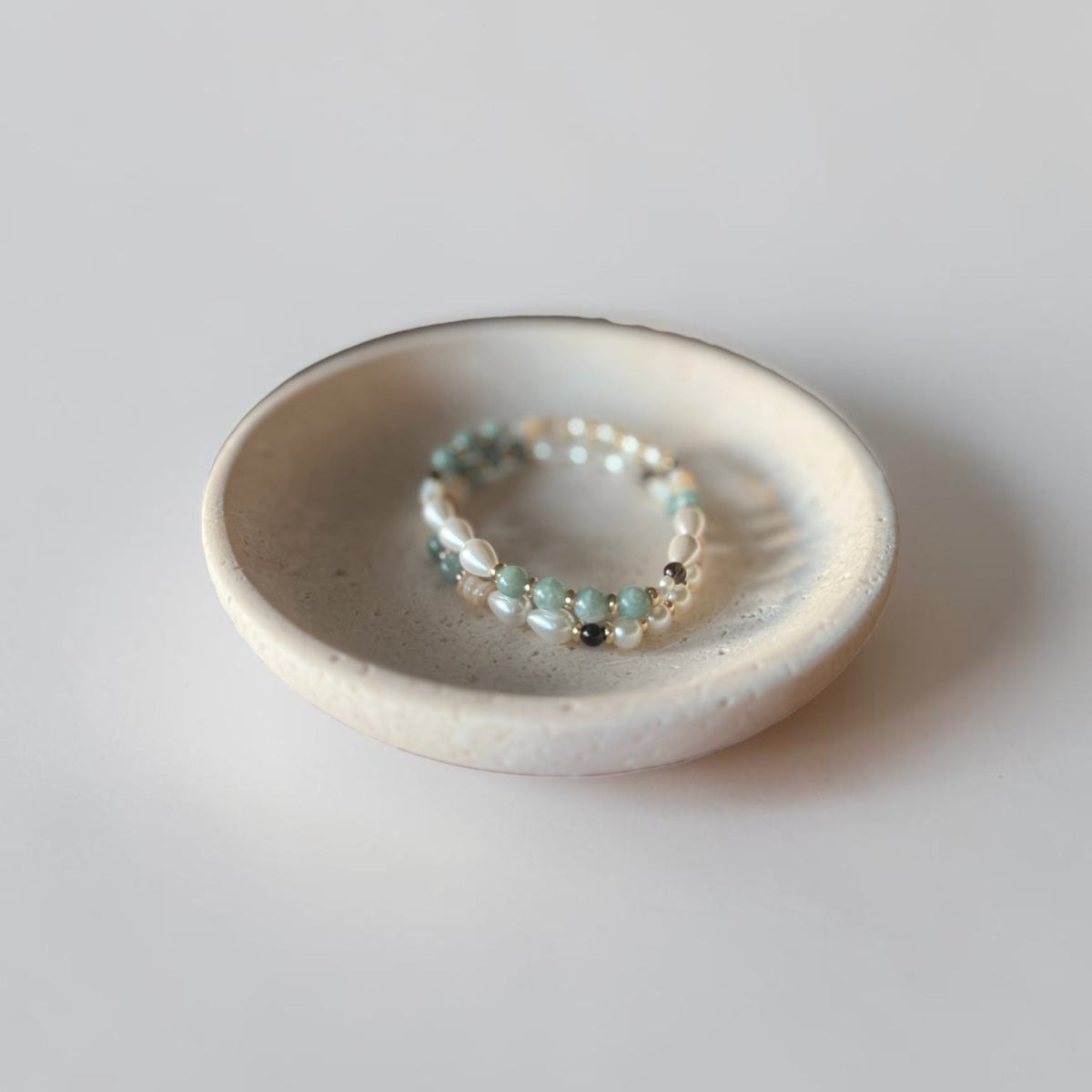 A delicate beaded bracelet with small light green, white, and silver beads resting inside a small, shallow, speckled off-white ceramic dish on a neutral light gray background.
