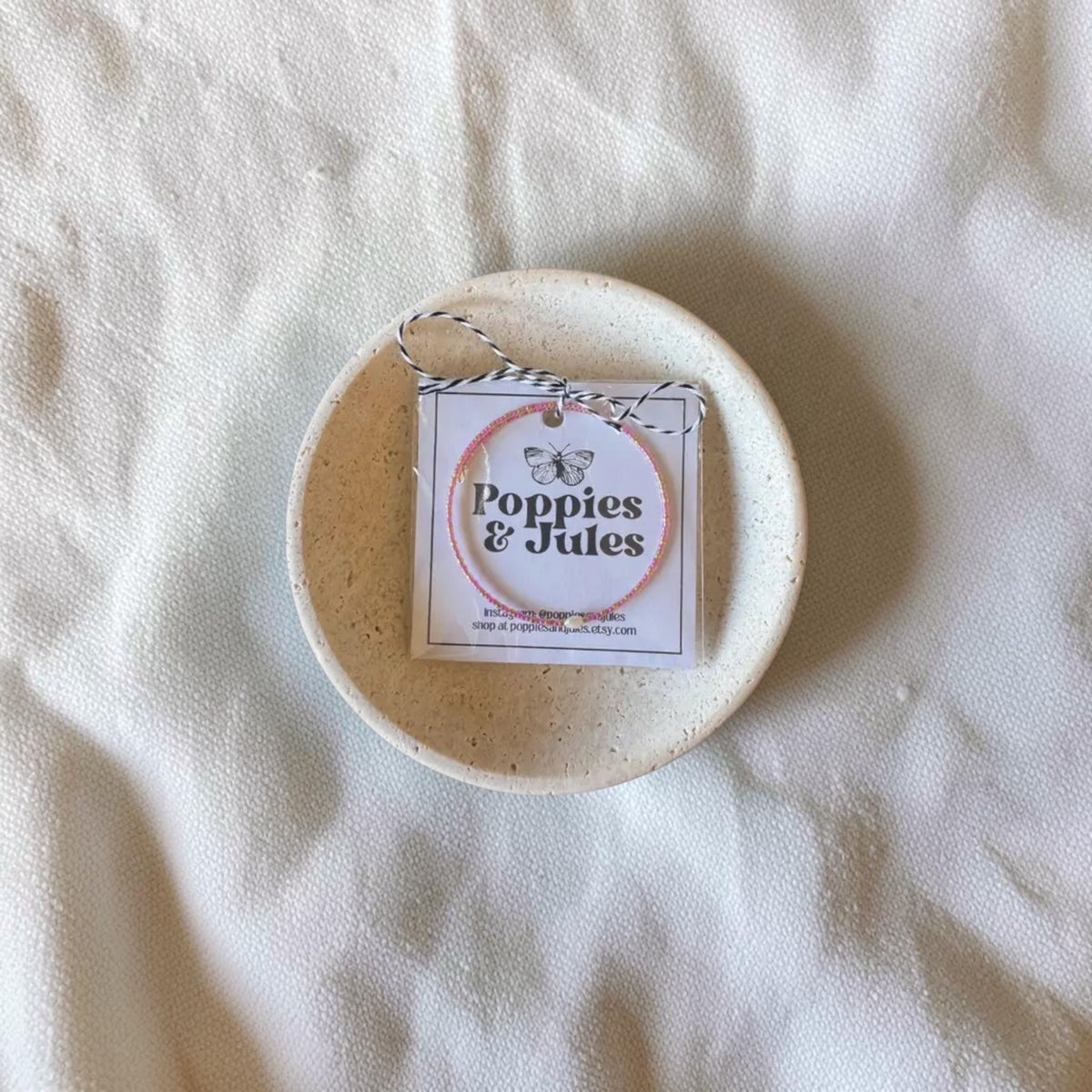 A small round ceramic dish holding a jewelry tin with a hanging tag that reads 'Poppies & Jules' on a textured off-white fabric background.