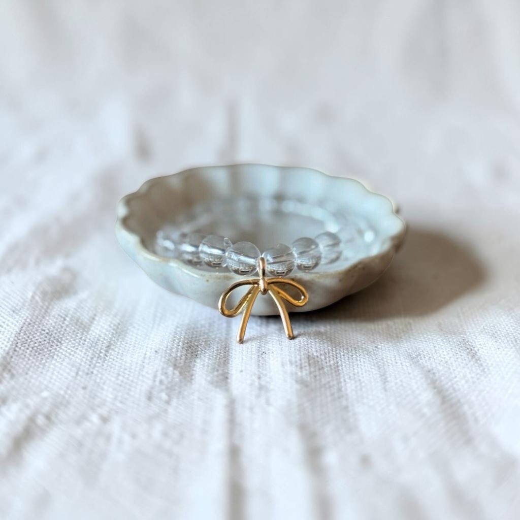 A small clear beaded bracelet with a gold bow charm, displayed inside a scalloped off-white ceramic dish on a textured white linen fabric background.
