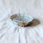 A small clear beaded bracelet with a gold bow charm, displayed inside a scalloped off-white ceramic dish on a textured white linen fabric background.