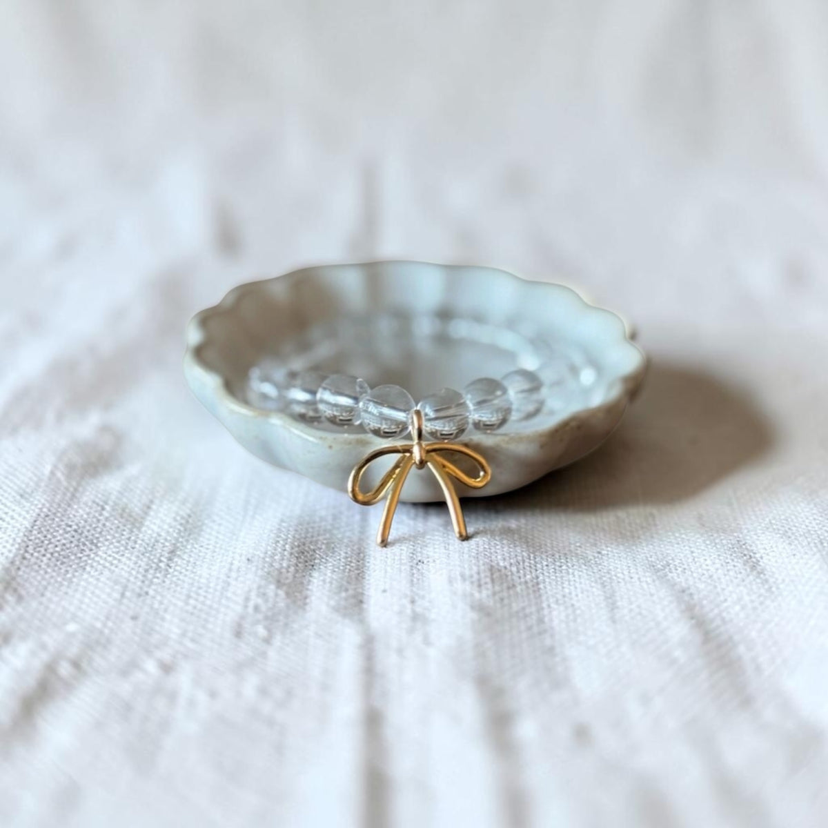 A small clear beaded bracelet with a gold bow charm, displayed inside a scalloped off-white ceramic dish on a textured white linen fabric background.