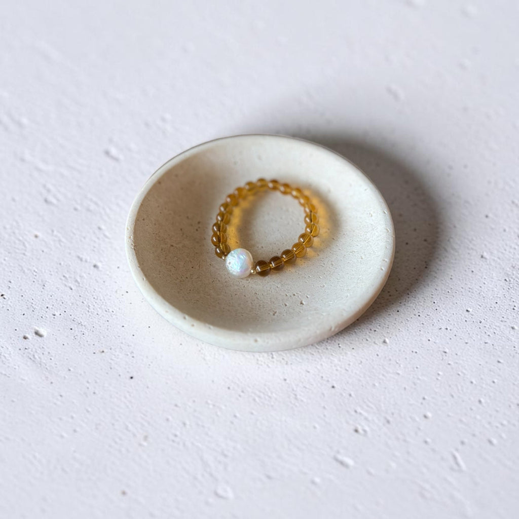 A delicate gold beaded ring with a single white pearl, displayed on a small round ceramic dish against a textured light gray background.
