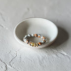 A beaded stretch ring with white, black, orange, and gold ceramic beads, displayed on a small white ceramic dish against a textured light gray background.