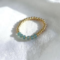 A delicate gold beaded bracelet featuring five small round light blue-green beads, resting on a textured white fabric background.