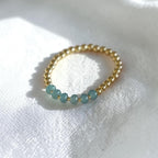 A delicate gold beaded bracelet featuring five small round light blue-green beads, resting on a textured white fabric background.