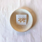 A pair of gold bow earrings with white pearl drops, displayed on a white card labeled 'Poppies + Jules' and placed on a round beige ceramic dish over a white fabric background.