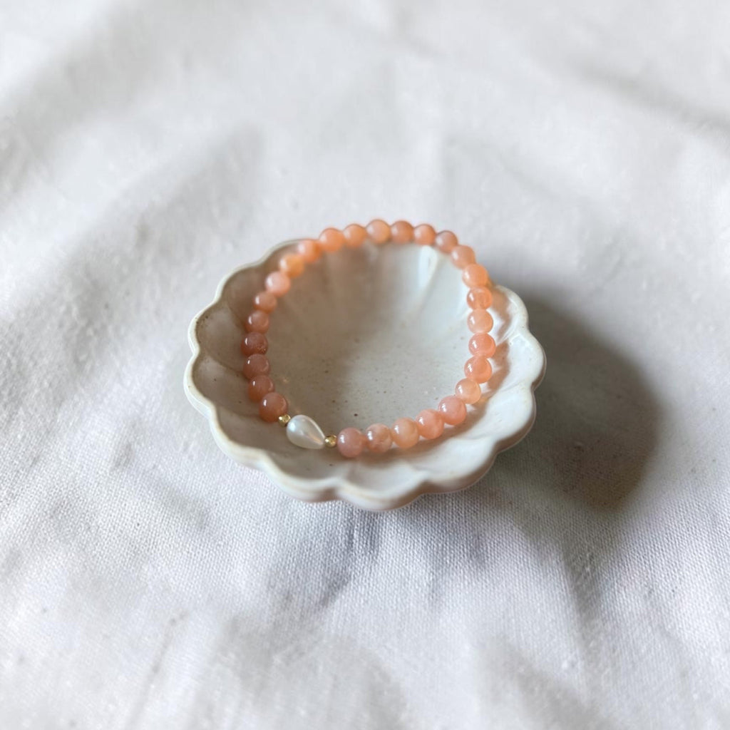 A peach-orange beaded bracelet with a small white pearl accent, displayed inside a small white ceramic dish on a textured white fabric background.