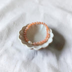 A peach-orange beaded bracelet with a small white pearl accent, displayed inside a small white ceramic dish on a textured white fabric background.