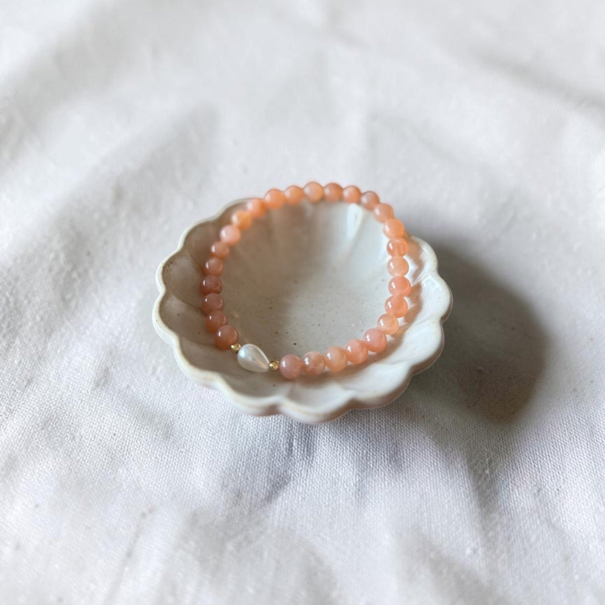 A peach-orange beaded bracelet with a small white pearl accent, displayed inside a small white ceramic dish on a textured white fabric background.