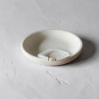 A small round ceramic dish holding a delicate beaded bracelet with pale blue beads and a gold clasp, set against a textured light gray background.