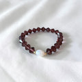 A beaded stretch bracelet featuring faceted dark burgundy beads and a central white iridescent pearl bead, displayed on a soft white fabric background.