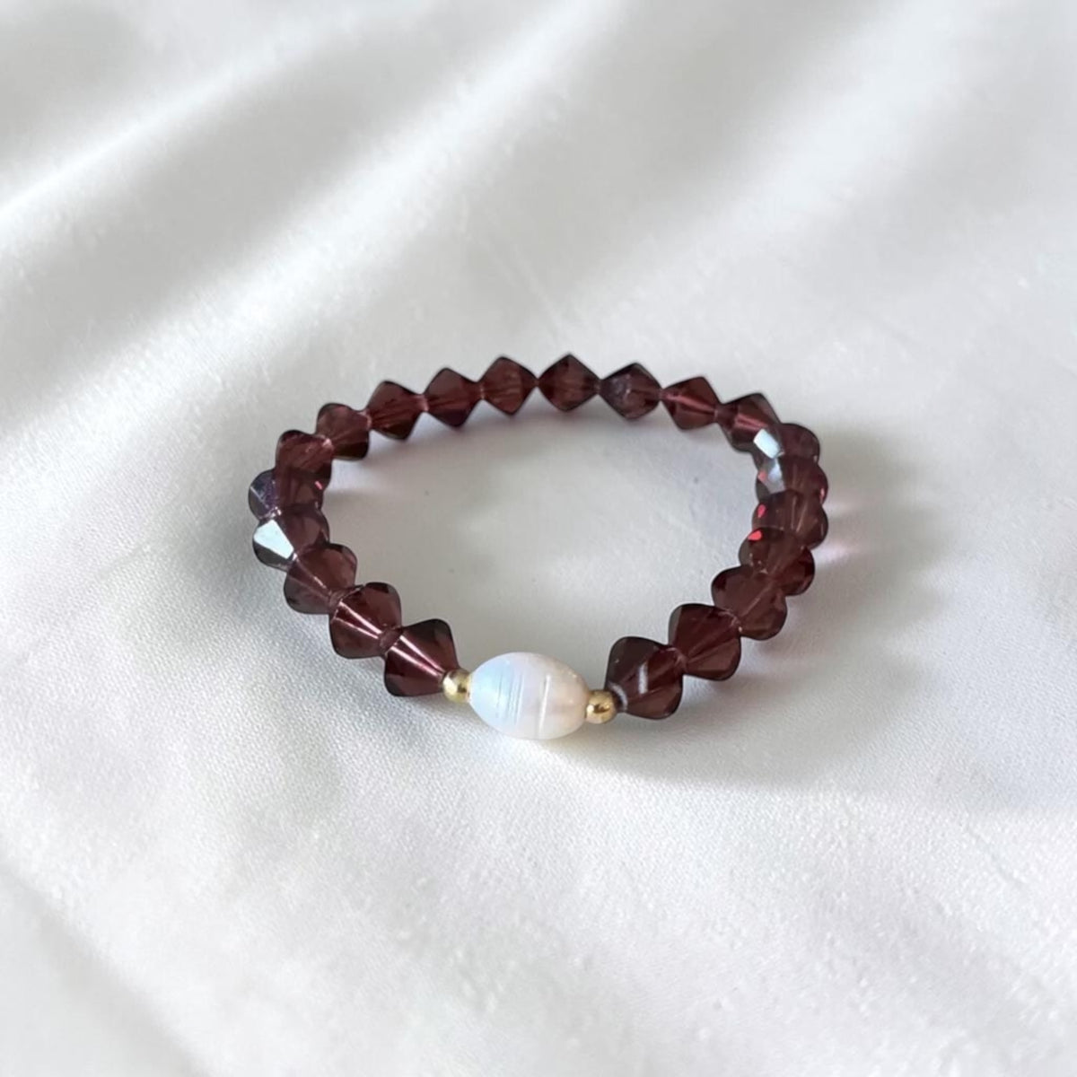 A beaded stretch bracelet featuring faceted dark burgundy beads and a central white iridescent pearl bead, displayed on a soft white fabric background.