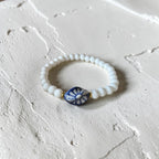 A beaded stretch bracelet with white round beads and a central blue and white floral accent bead, displayed on a textured off-white surface.