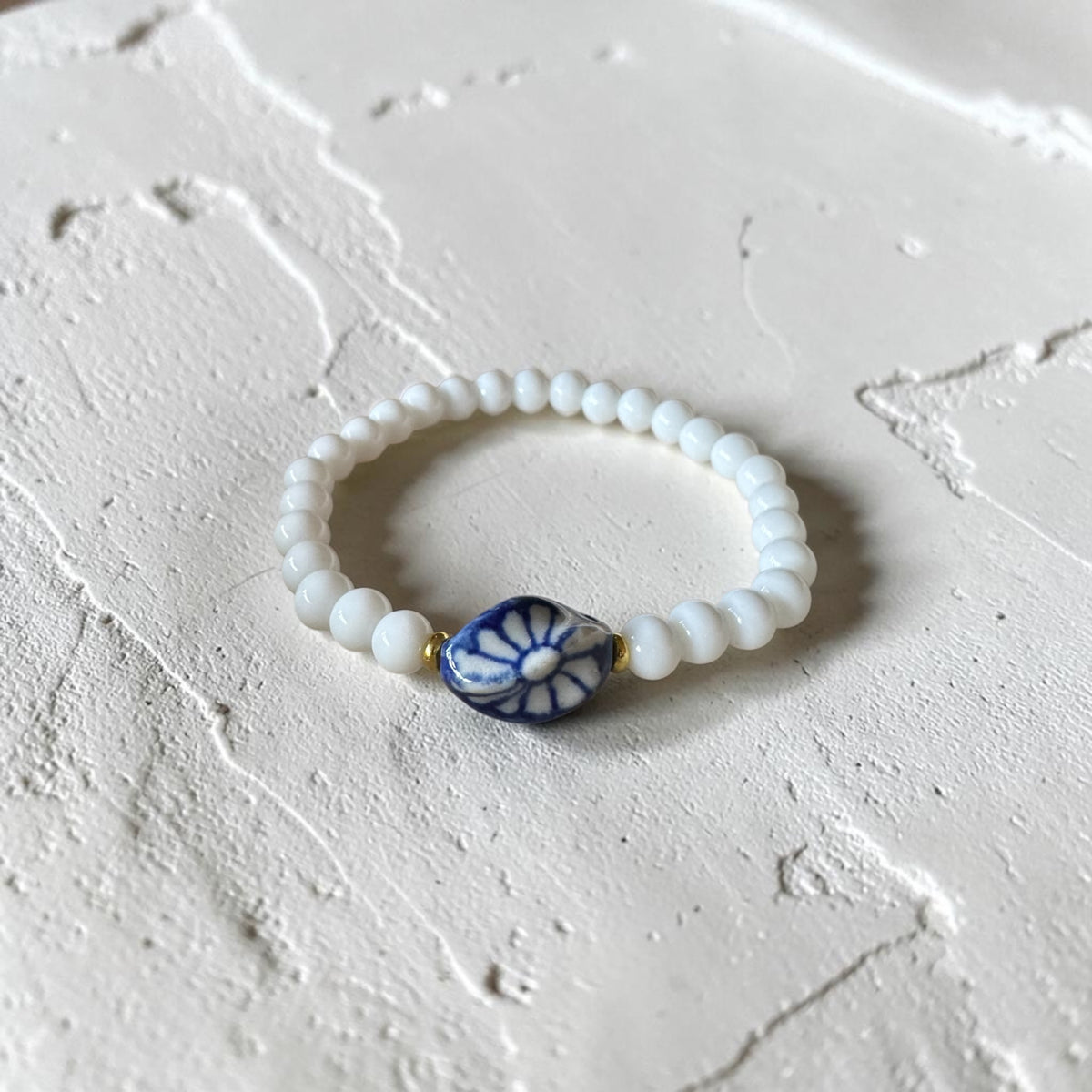A beaded stretch bracelet with white round beads and a central blue and white floral accent bead, displayed on a textured off-white surface.