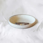 A beaded stretch bracelet with dark brown beads and a small gold-toned accent, displayed inside a small, round, textured ceramic dish on a soft white fabric background.