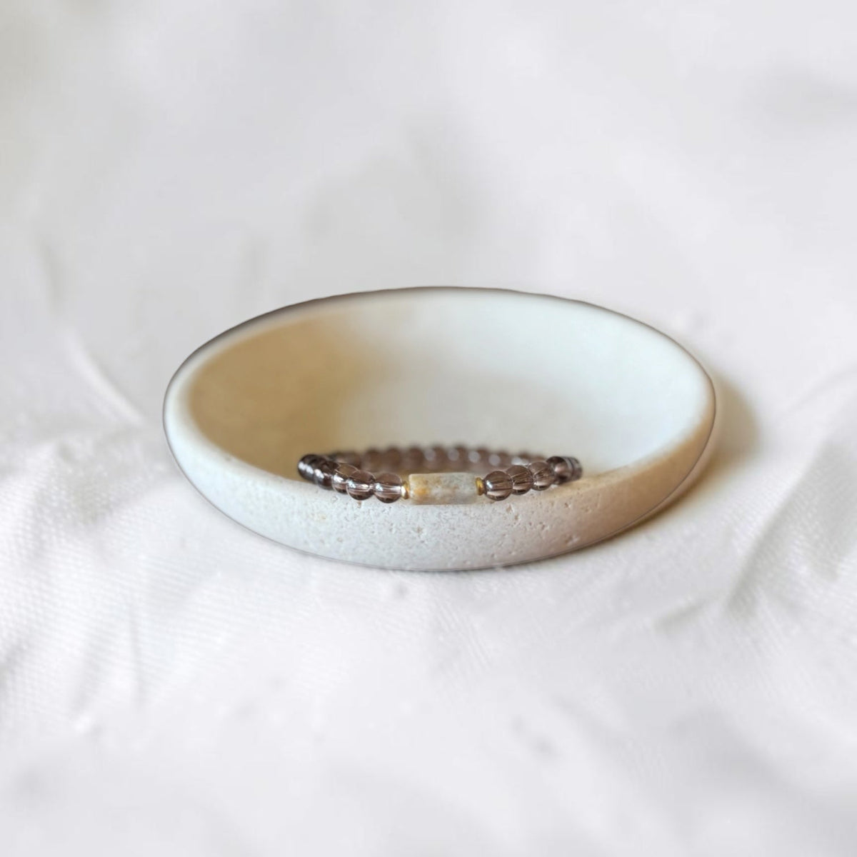 A beaded stretch bracelet with dark brown beads and a small gold-toned accent, displayed inside a small, round, textured ceramic dish on a soft white fabric background.