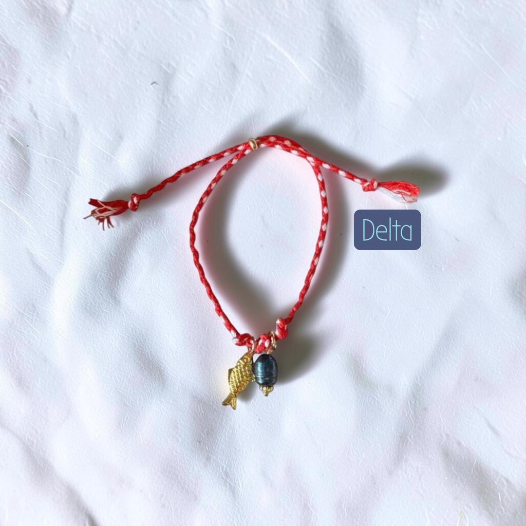 A red braided cord bracelet with a small gold fish charm and a dark gray bead, displayed on a white fabric background with a blue 'Delta' label.