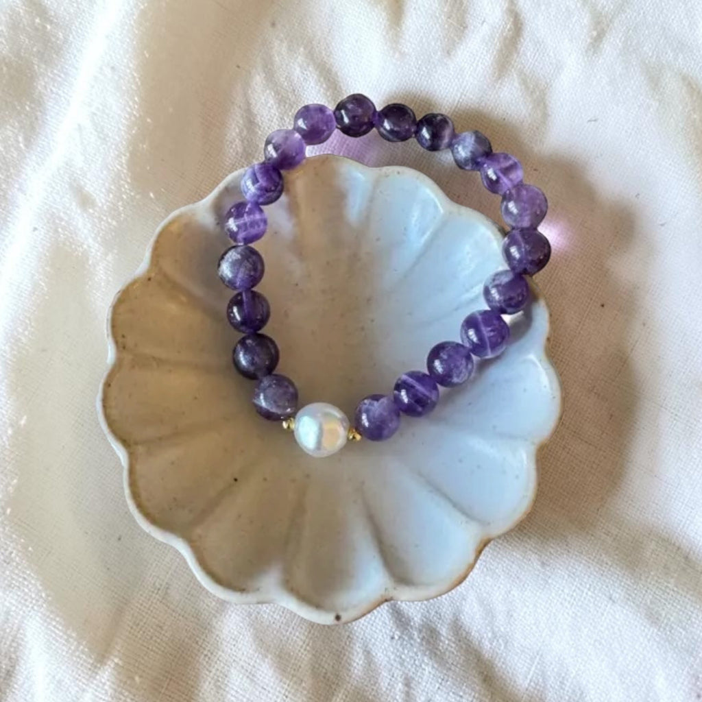 A beaded stretch bracelet made of purple amethyst beads with a white pearl accent, displayed on a small scalloped ceramic dish over a white fabric background.