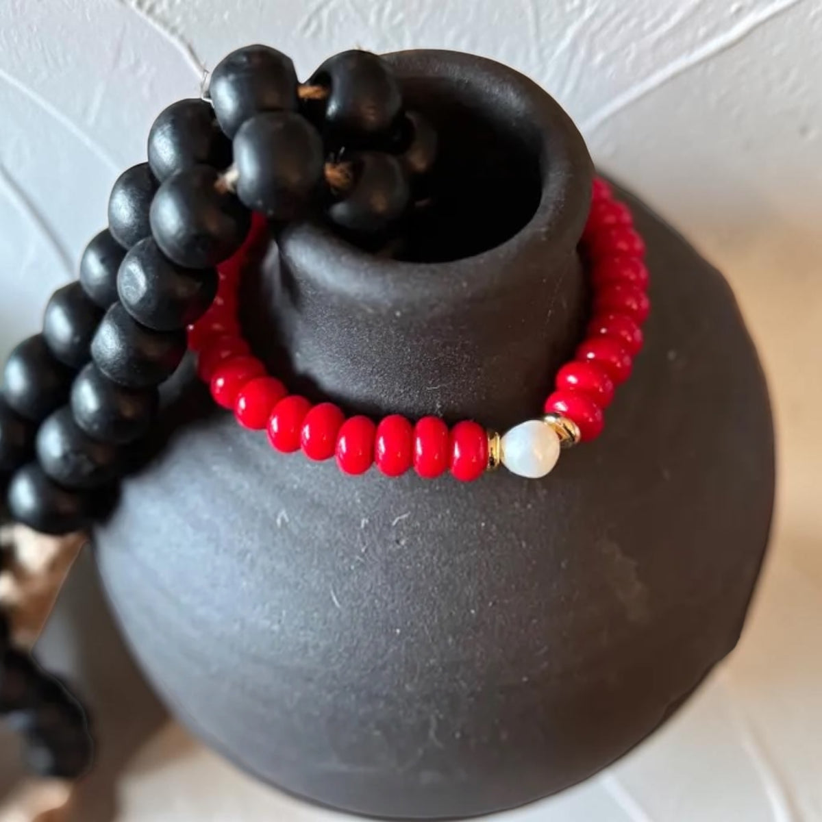 A red beaded bracelet with a white bead and gold spacer, displayed on a matte black ceramic vase next to a black beaded necklace.