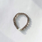 A beaded stretch bracelet made of translucent smoky brown beads with a single white freshwater pearl accent, resting on a white textured surface with soft shadows.