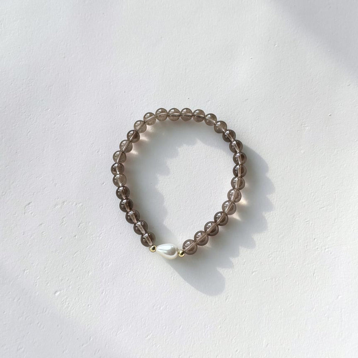 A beaded stretch bracelet made of translucent smoky brown beads with a single white freshwater pearl accent, resting on a white textured surface with soft shadows.