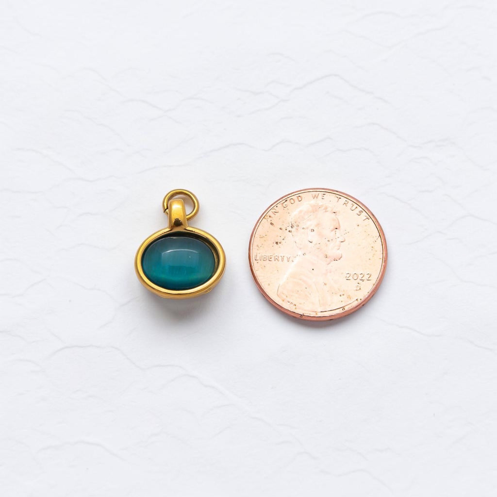 The bracelet charm with jump hoop. Outlined in gold and a stone inside. Sitting next to a penny on white texture background. Showing the comparison of the size. 