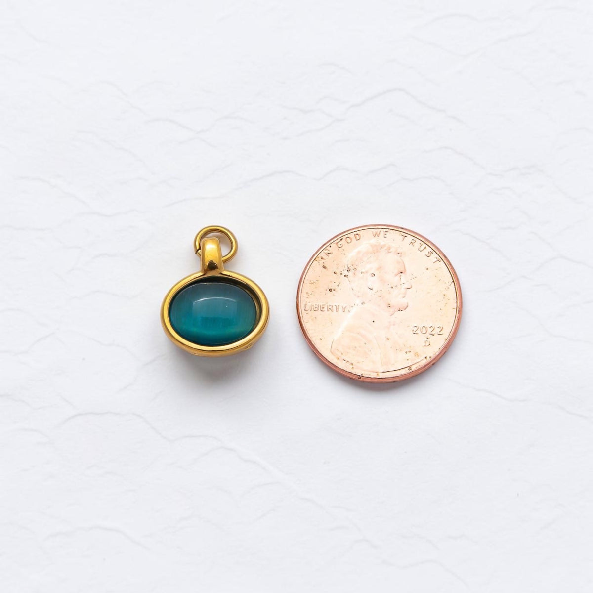 The bracelet charm with jump hoop. Outlined in gold and a stone inside. Sitting next to a penny on white texture background. Showing the comparison of the size. 