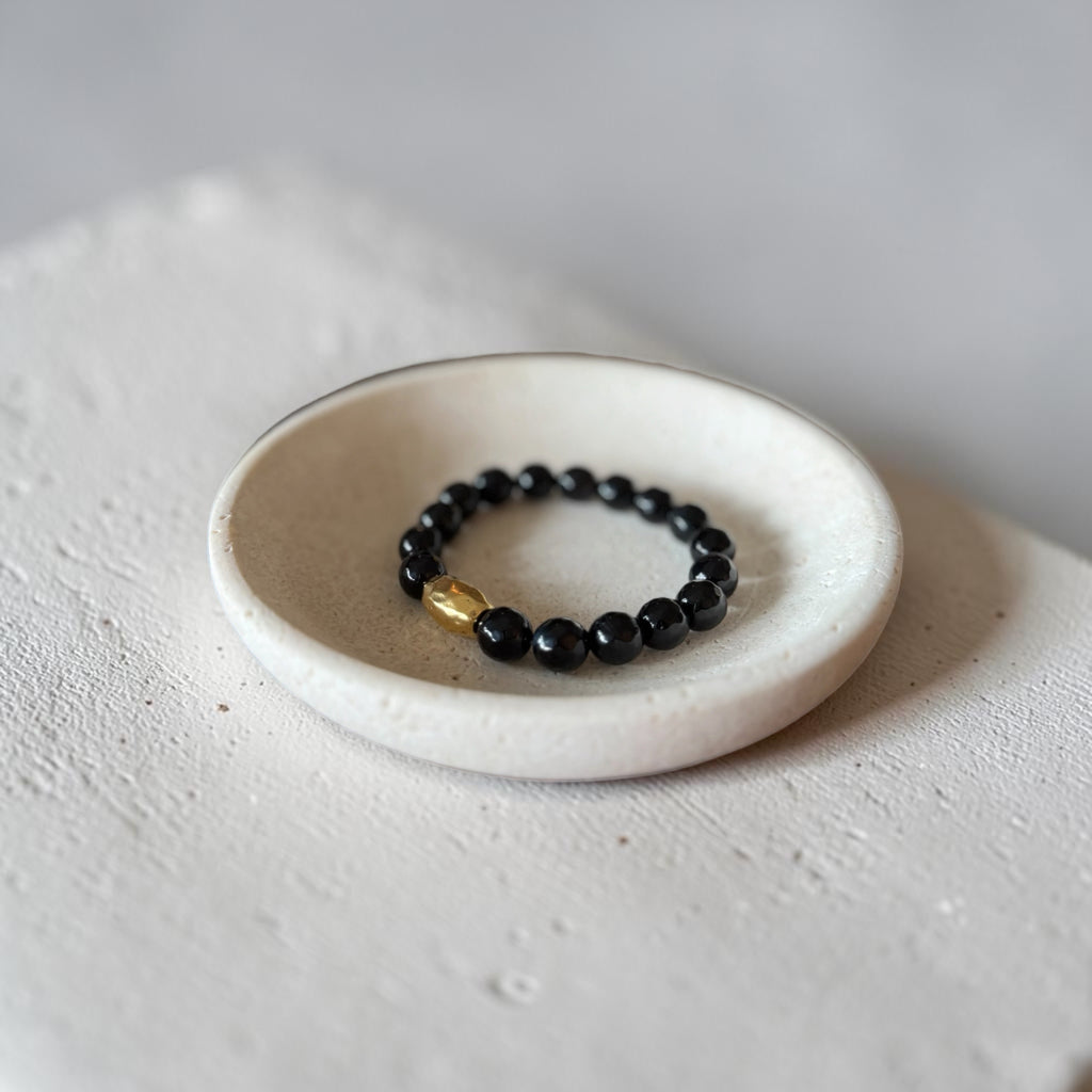 A beaded stretch bracelet with glossy black round beads and a small gold accent bead, displayed inside a small round off-white ceramic dish on a textured light surface.