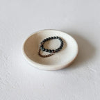 A beaded stretch bracelet with dark gray beads and a bronze chain extender, displayed in a small, round, textured off-white ceramic dish on a neutral light gray background.