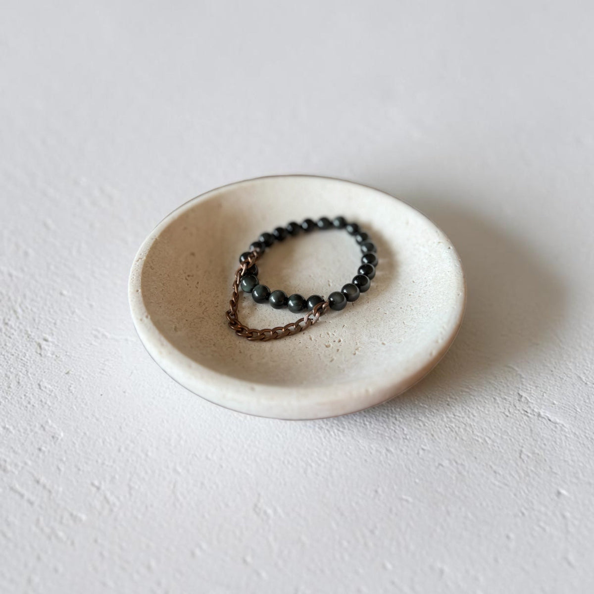 A beaded stretch bracelet with dark gray beads and a bronze chain extender, displayed in a small, round, textured off-white ceramic dish on a neutral light gray background.