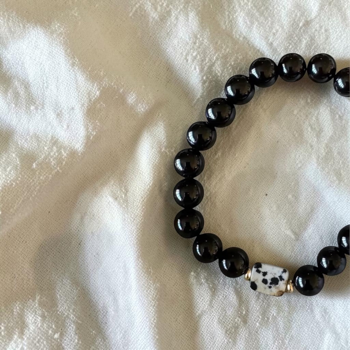 A beaded bracelet made of glossy black round beads with a white cylindrical bead featuring black spots, resting on a textured white fabric background.