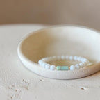 A beaded stretch bracelet with white round beads and a central light green rectangular accent bead, displayed inside a shallow cream-colored ceramic dish on a textured beige surface.