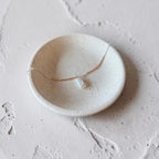 A delicate gold necklace with a small white pearl pendant resting inside a small, round, speckled ceramic dish on a textured light gray surface.