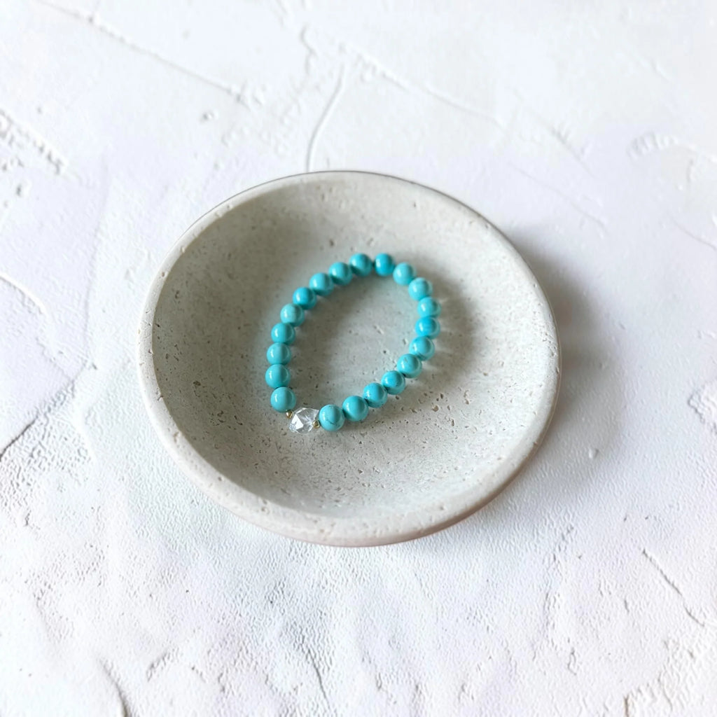 A turquoise beaded bracelet with a small silver charm, displayed inside a small speckled ceramic dish on a textured white surface.